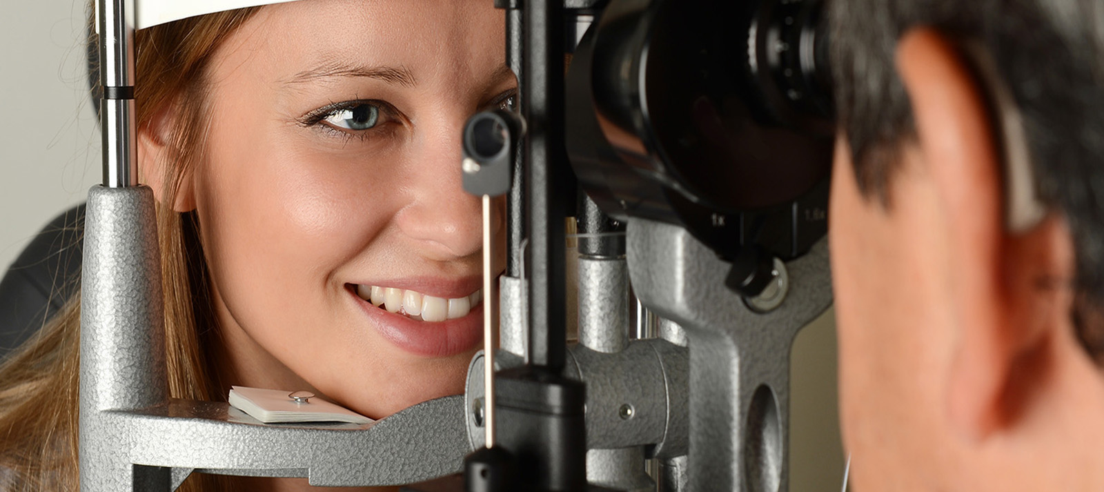 The image shows a woman sitting in an eye exam chair with her eyes open while looking at a screen, and a man standing behind her, adjusting the equipment of the eye machine.