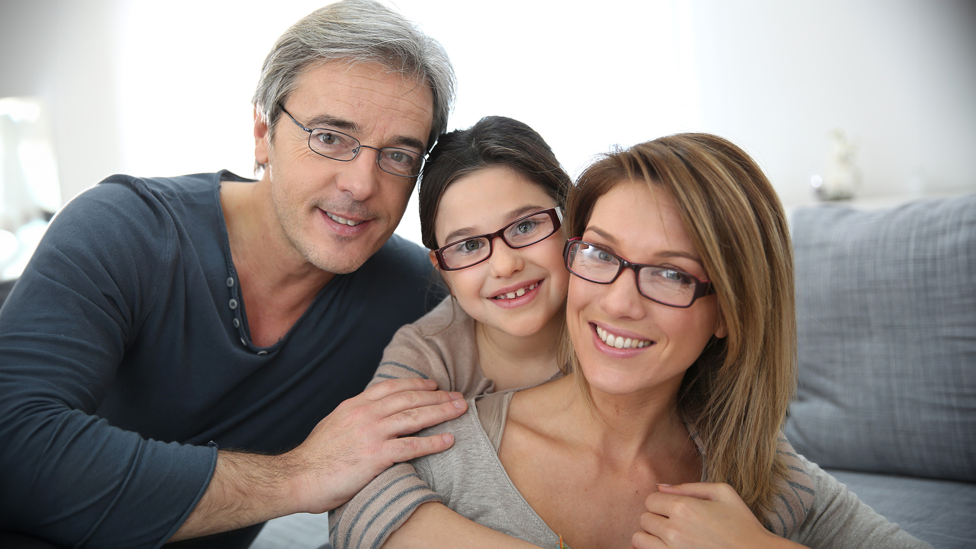 A family of three posing together with smiles, the father standing behind the mother and child, all appearing happy and content.
