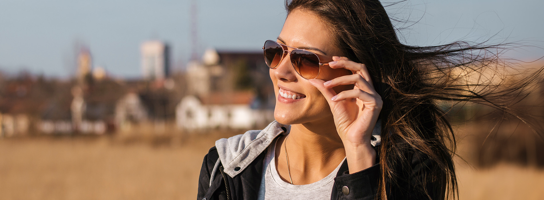 The image shows a woman with sunglasses on her head, smiling at the camera, standing outdoors during daylight.