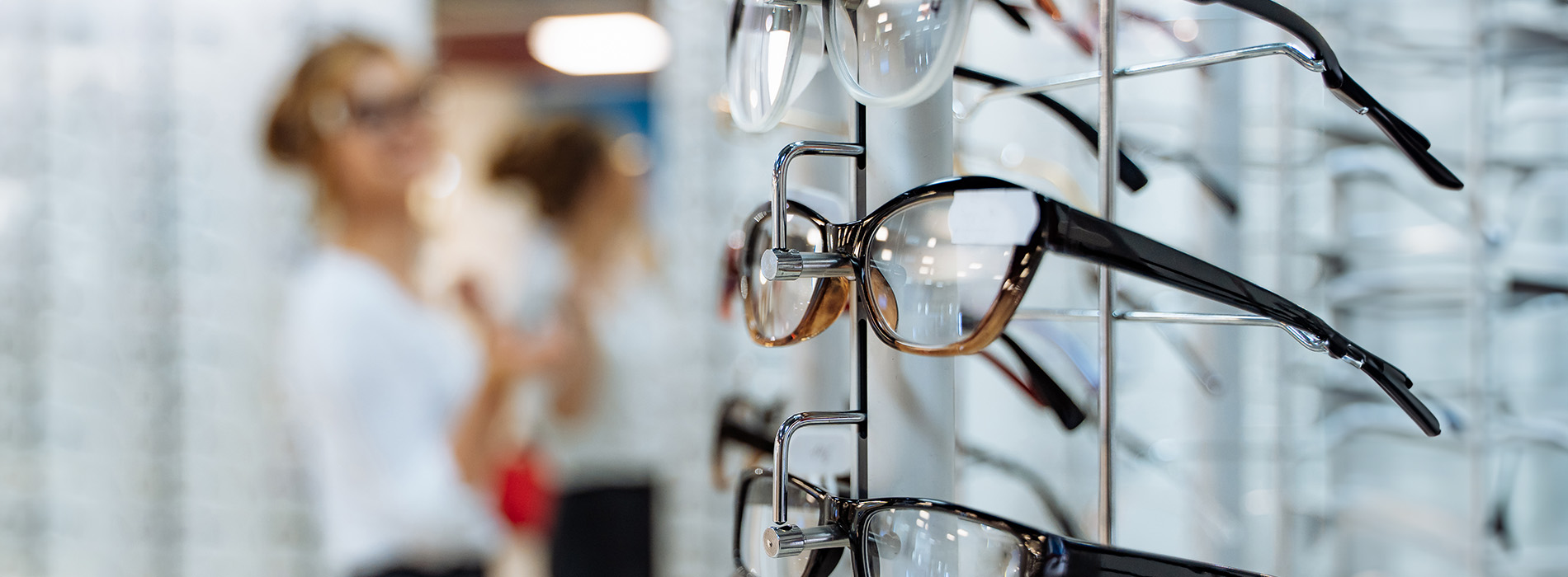 The image shows a collection of eyeglasses displayed on a rack with people standing nearby, suggesting this is likely an optical store.