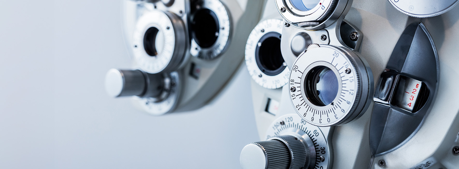 The image shows a collection of eye glasses lenses arranged vertically with various sizes and designs, set against a blurred background that includes an optical device with dials and markings, which suggests a focus on optics or vision-related equipment.