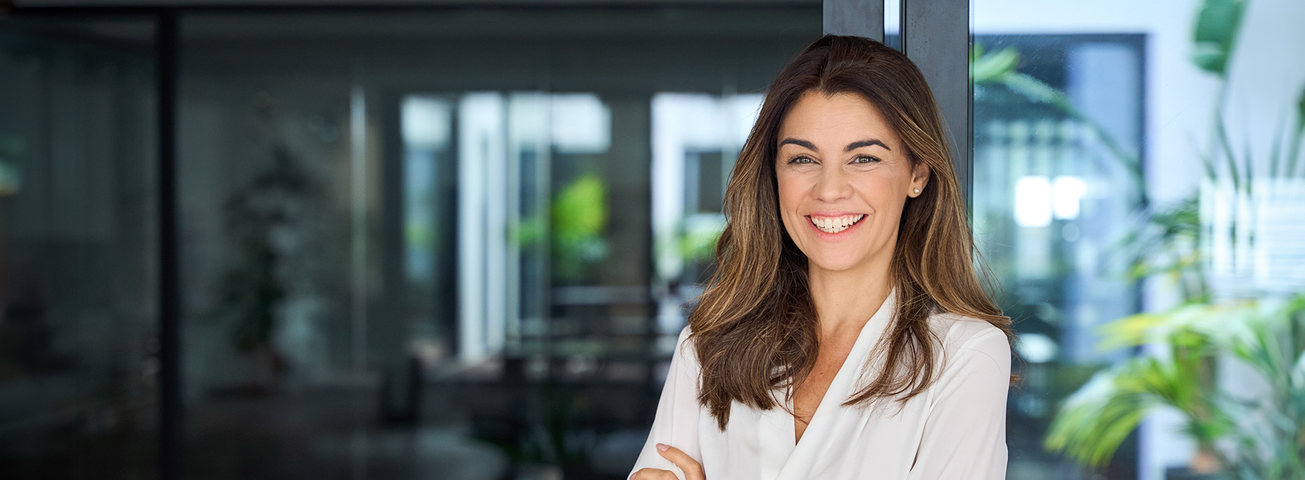 The image shows a woman smiling at the camera, dressed in professional attire, standing in an office environment with a blurred background.