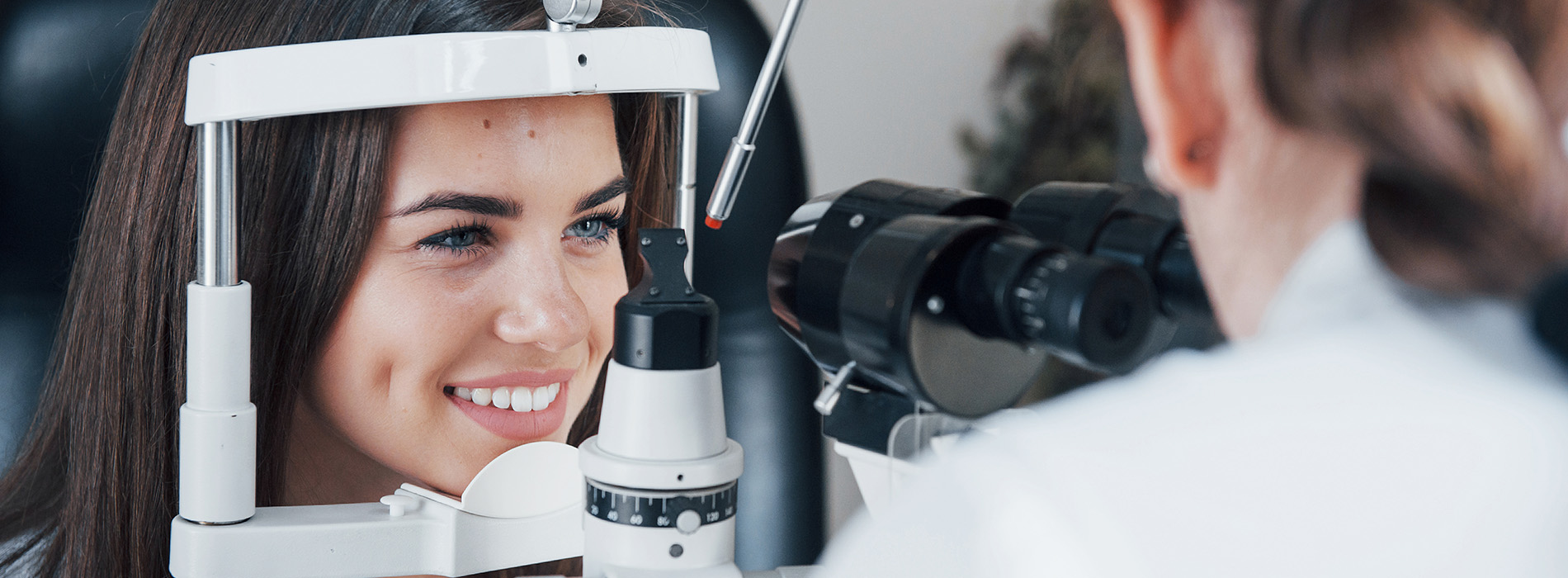 The image shows a woman sitting at an eye exam station with an optometrist s chair and equipment, smiling towards the camera.