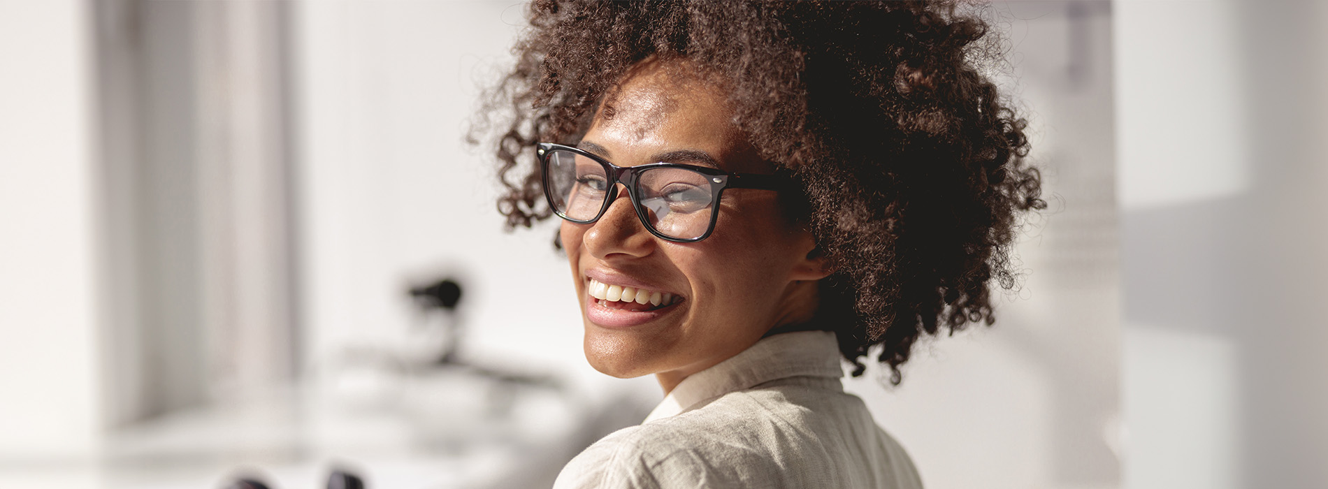 The image features a woman with curly hair smiling and looking over her shoulder, wearing glasses and a dark top, against a blurred background.