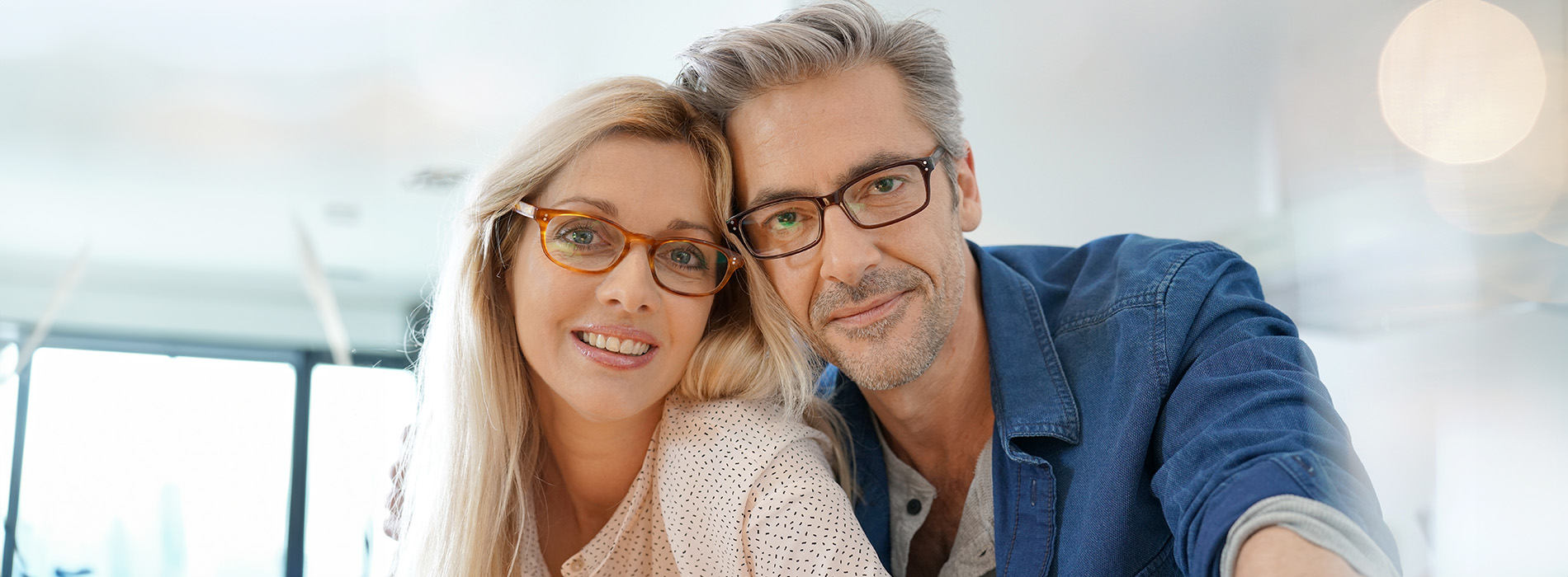 The image features a man and woman posing together with a warm smile, both wearing glasses and casual attire  they are seated indoors, likely in a restaurant or caf   setting.