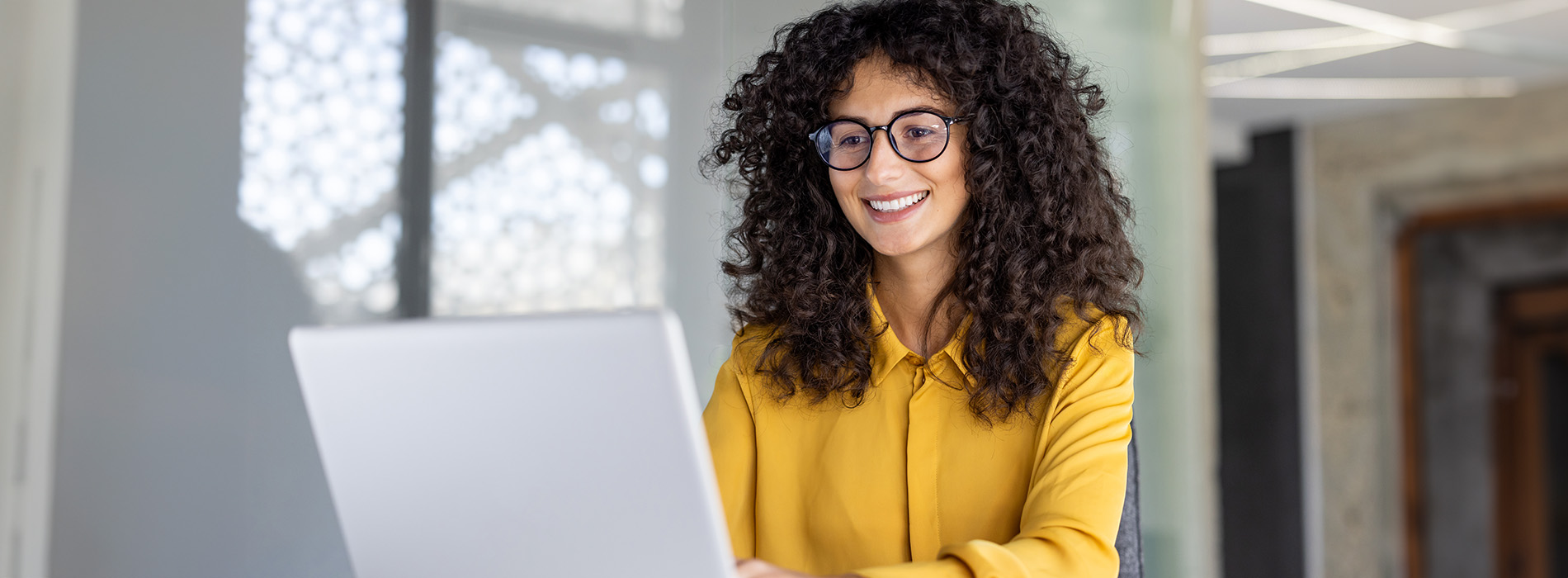 The image shows a woman with curly hair seated at a desk, using a laptop computer, wearing glasses and a yellow top, set against an office environment with modern decor.