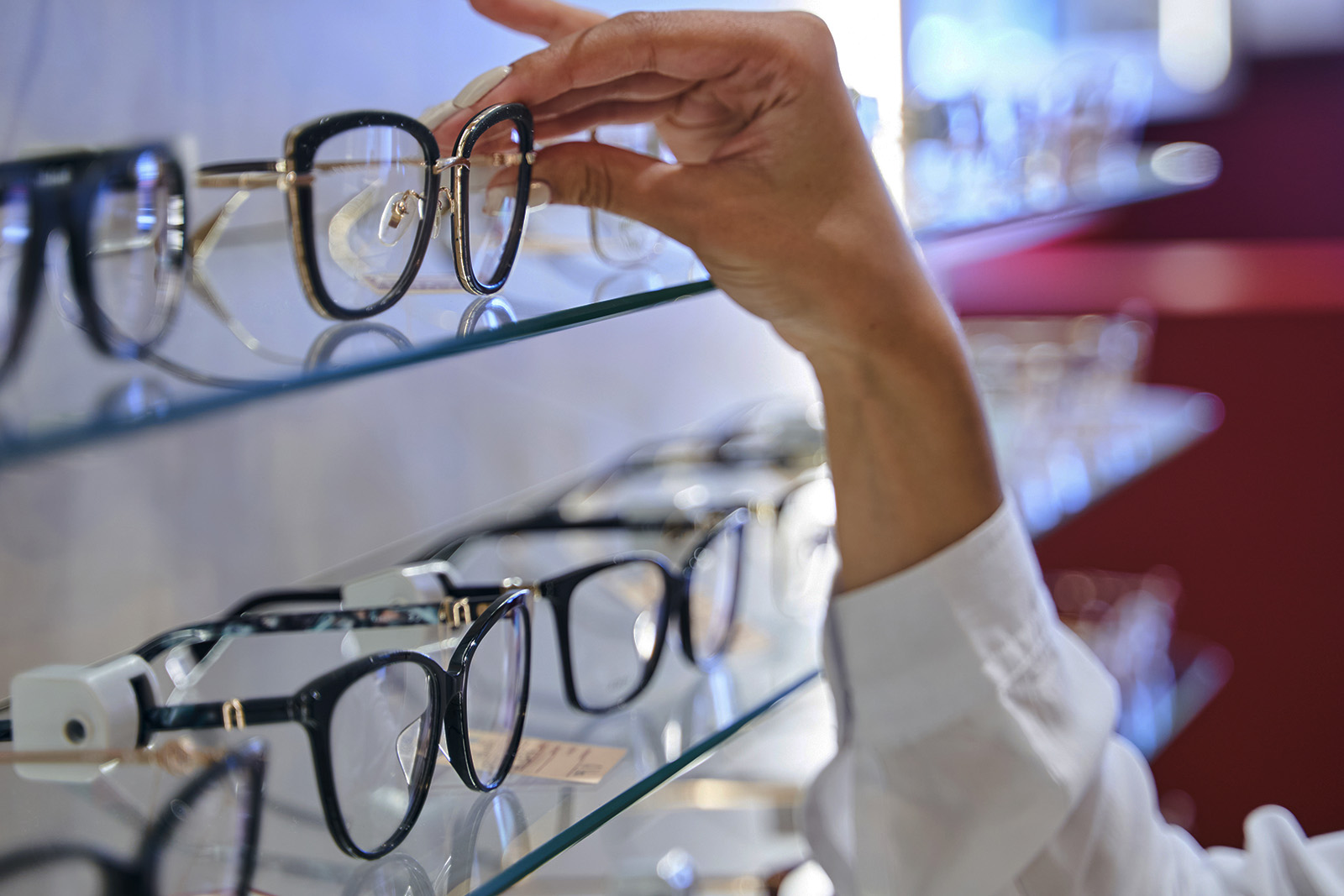 The image shows a person s hand reaching out to display a pair of eyeglasses on a shelf in an optical store, with multiple pairs of glasses visible in the background.