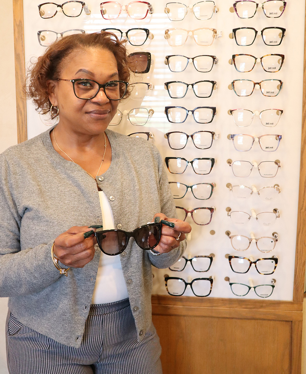 A woman wearing glasses stands next to a display case filled with various pairs of eyeglasses.