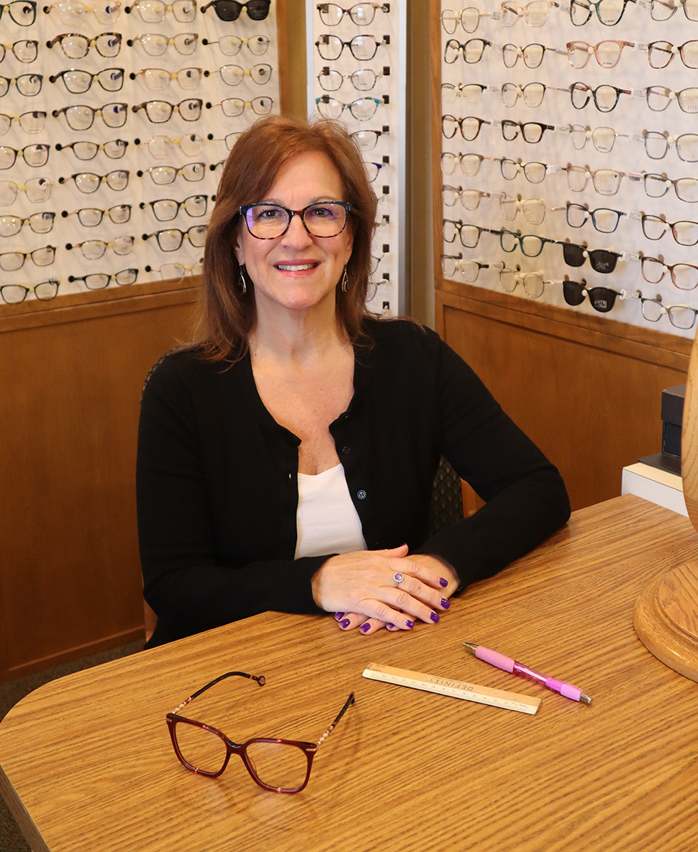 The image shows a woman sitting at a desk with eyeglasses on display behind her.