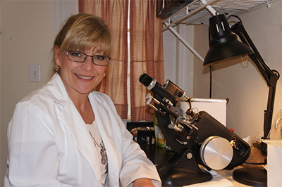 The image shows a woman standing behind a microscope, wearing glasses and a white lab coat, with a desk lamp illuminating the workspace.