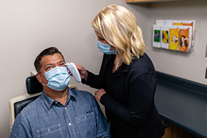 A woman wearing a face mask is assisting a man with a medical device, possibly during a dental examination or procedure.