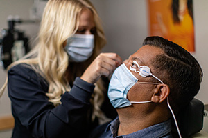 A woman wearing a surgical mask is carefully adjusting a man s eyeglasses while he sits in a chair with his eyes closed.