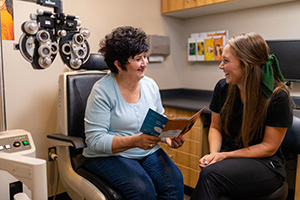The image shows two individuals, an older woman and a younger woman, sitting together in an optometrist s office, with the older woman wearing glasses and holding a booklet, while the younger woman is seated across from her, smiling and looking at her.