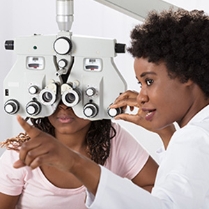 The image shows two individuals engaged in an eye examination, with one person seated behind an optical device, likely a slit lamp, while another stands beside them pointing towards a specific area of interest within the device s viewfield.