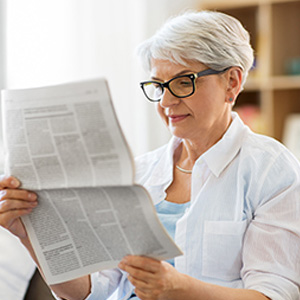 An elderly woman reading a newspaper with glasses on her face.