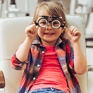 A young child sitting in an eye exam chair with oversized glasses on their face, smiling at the camera.