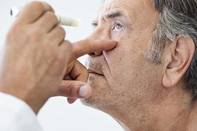 The image shows a man sitting with his eyes closed while another person administers something to him near his eye, likely related to an eye exam or treatment.