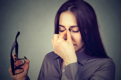 A woman with her eyes closed, holding a pair of glasses to her nose, with a contemplative expression.
