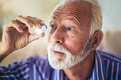 This is a color photograph featuring an older man with white hair and a beard, wearing a blue shirt, looking through a magnifying glass at a small object held up to his eye. The background is out of focus but suggests an indoor setting with warm lighting.