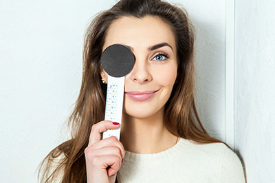 The image shows a woman holding up a ruler with her face partially obscured by the ruler s edge, standing against a white background with a blurred pattern that could be interpreted as a shower curtain. She has long hair and appears to be indoors.