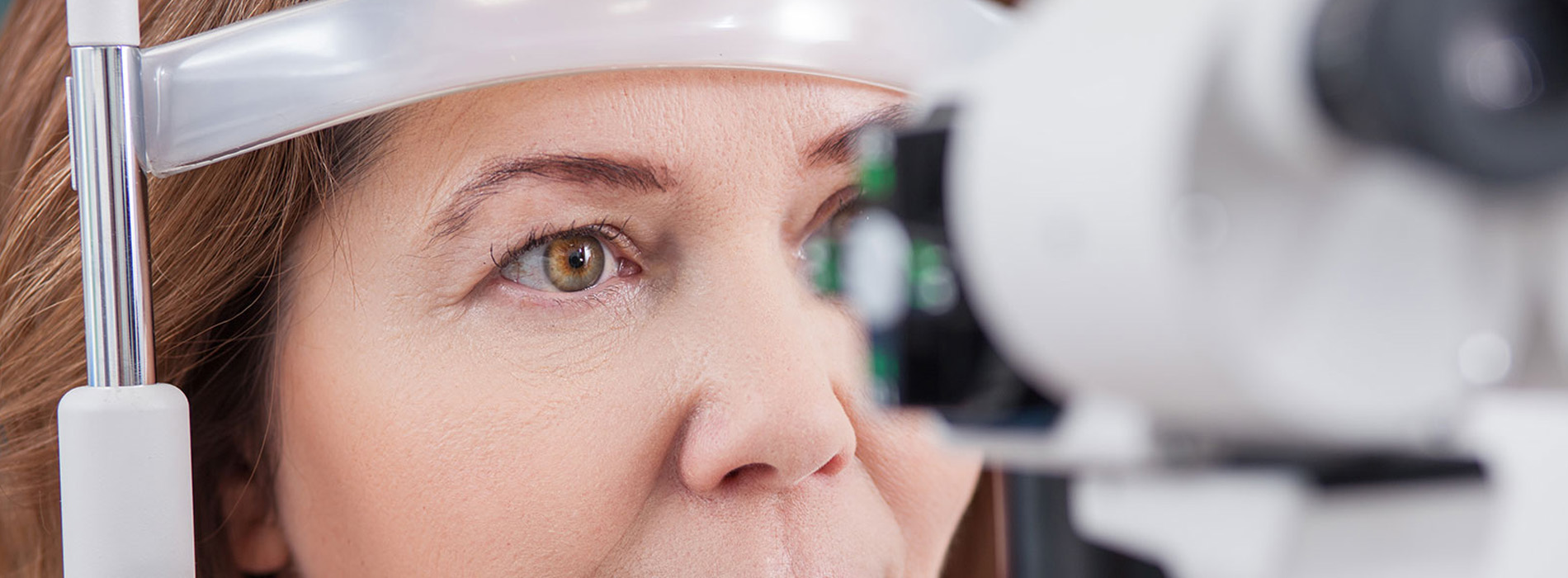 The image shows a woman with dark hair seated in front of an eye exam device, likely at an optometrist s office, wearing glasses on her face while looking directly forward.