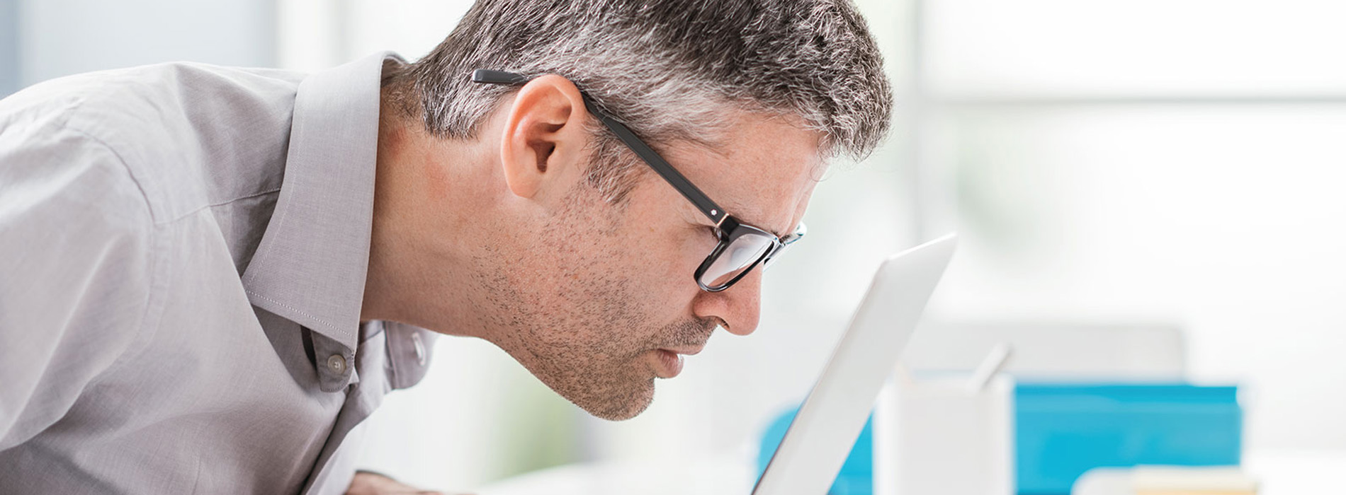 The image shows a man in a business setting, leaning over a table with papers, wearing glasses, and looking at something on the table while another person appears to be taking his picture from behind.
