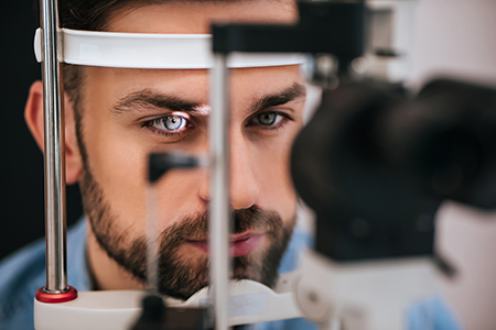 A man with blue eyes looking through an ophthalmoscope at his own eye, wearing a white headband, with medical equipment behind him.