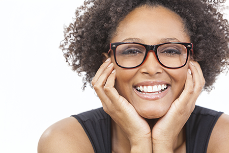The image shows a smiling woman with curly hair wearing glasses and a black top, leaning on her hand with her head tilted slightly towards the viewer.