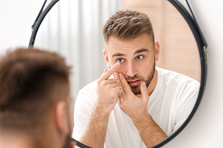 A man with short hair is looking at his reflection in a mirror while applying something to his face  he has a beard and mustache, and appears to be in a bathroom or grooming area.
