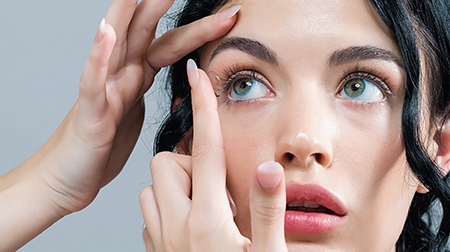 A woman with blue eyes looking into a mirror while applying makeup on her face.